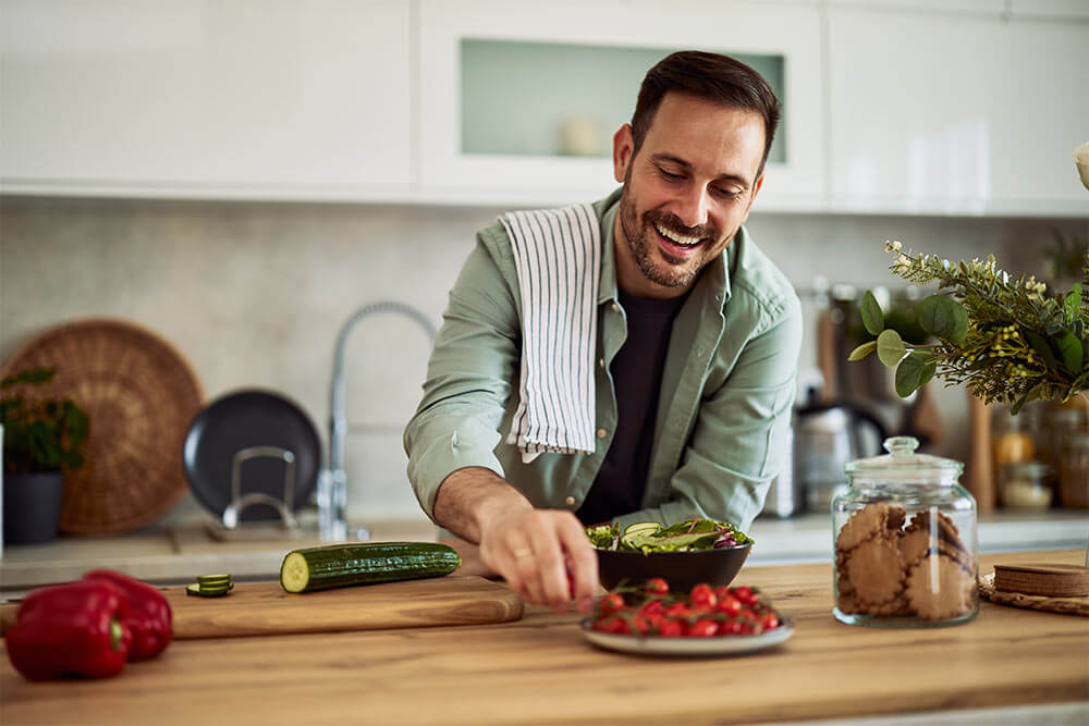 A smiling adult man taking a cherry tomato to mix it with the fresh and healthy vegetable salad